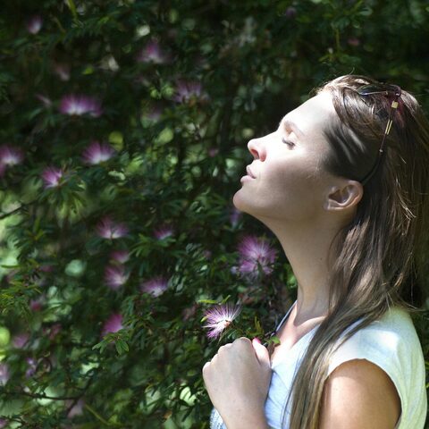 A woman is standing next to a bush taking in a deep breath with her eyes closed. 