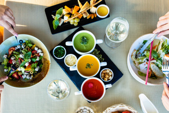 an overhead view of 2 people at a table. each has a different salad in front of them and there is a plate of sauces and another of cheese. 