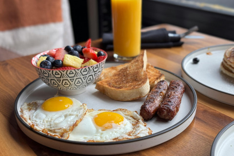 A plate of breakfast on a table. the plate has eggs, sausage, toast and a bowl of fruit. There is also a glass of orange juice on the table. 
