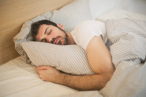 A man in a white shirt is asleep in a bed and cuddling a pillow