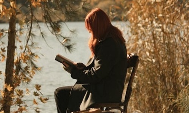 A woman with red hair is sitting on a bench near a lake reading a book
