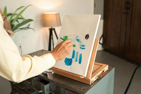 a persons arm and hand is shown in the picture. they are painting blue strokes on a canvas that is on an easel on a table