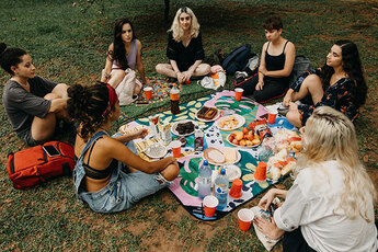 A group of women sitting in a circle on a blanket in a park. There is food and drinks and they are talking 
