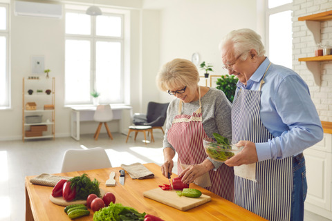 a happy, elderly man and woman are in the kitchen. they are preparing food and the woman is cutting vegetables 